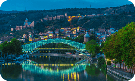 Illuminated bridge over the tranquil waters of Tbilisi's river at dusk.