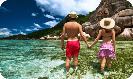 A couple hand in hand wading through Seychelles' clear waters with rocky shores in the distance.